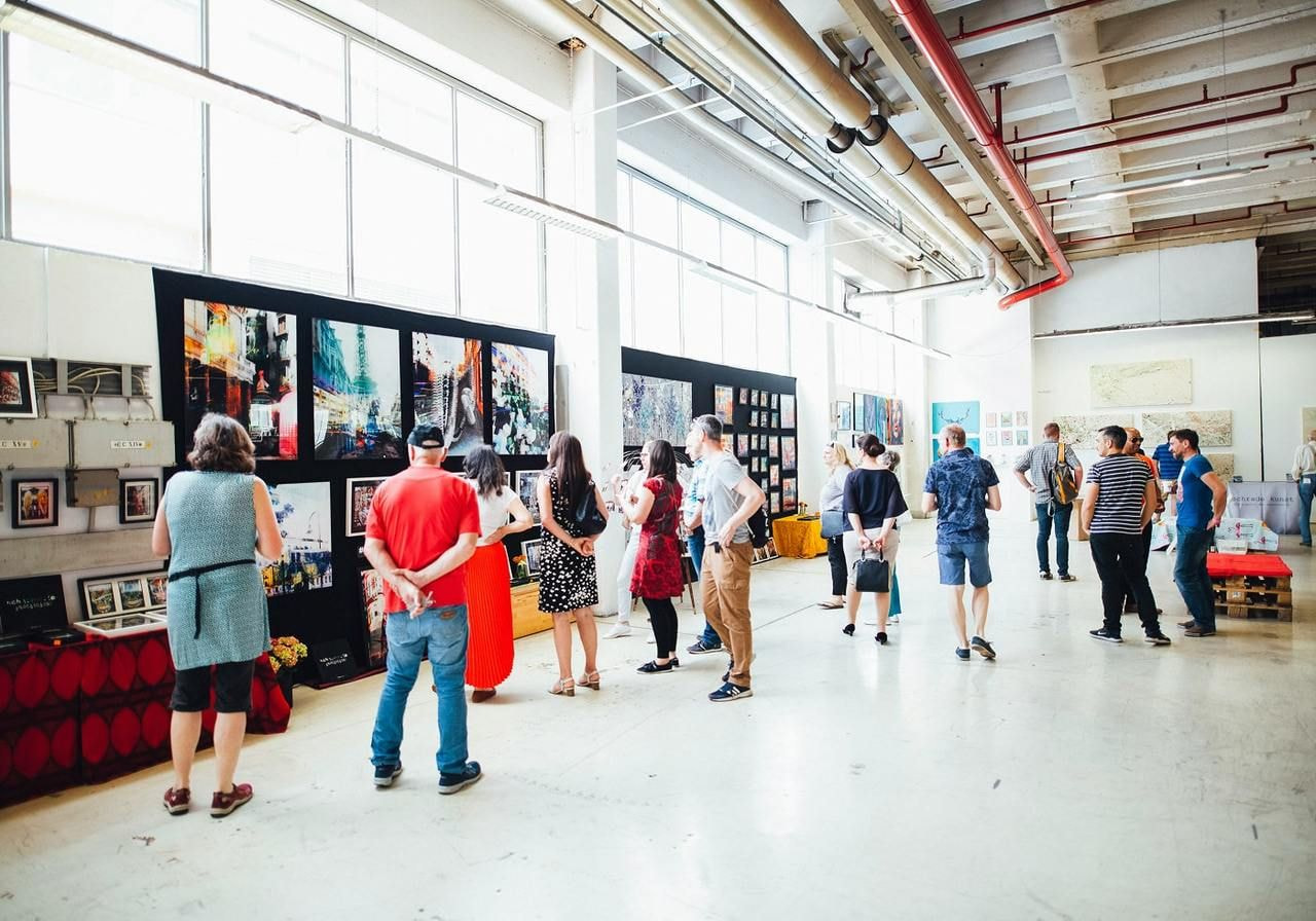 A group of people stands in an art fair, surrounded by framed pictures displayed on the walls.