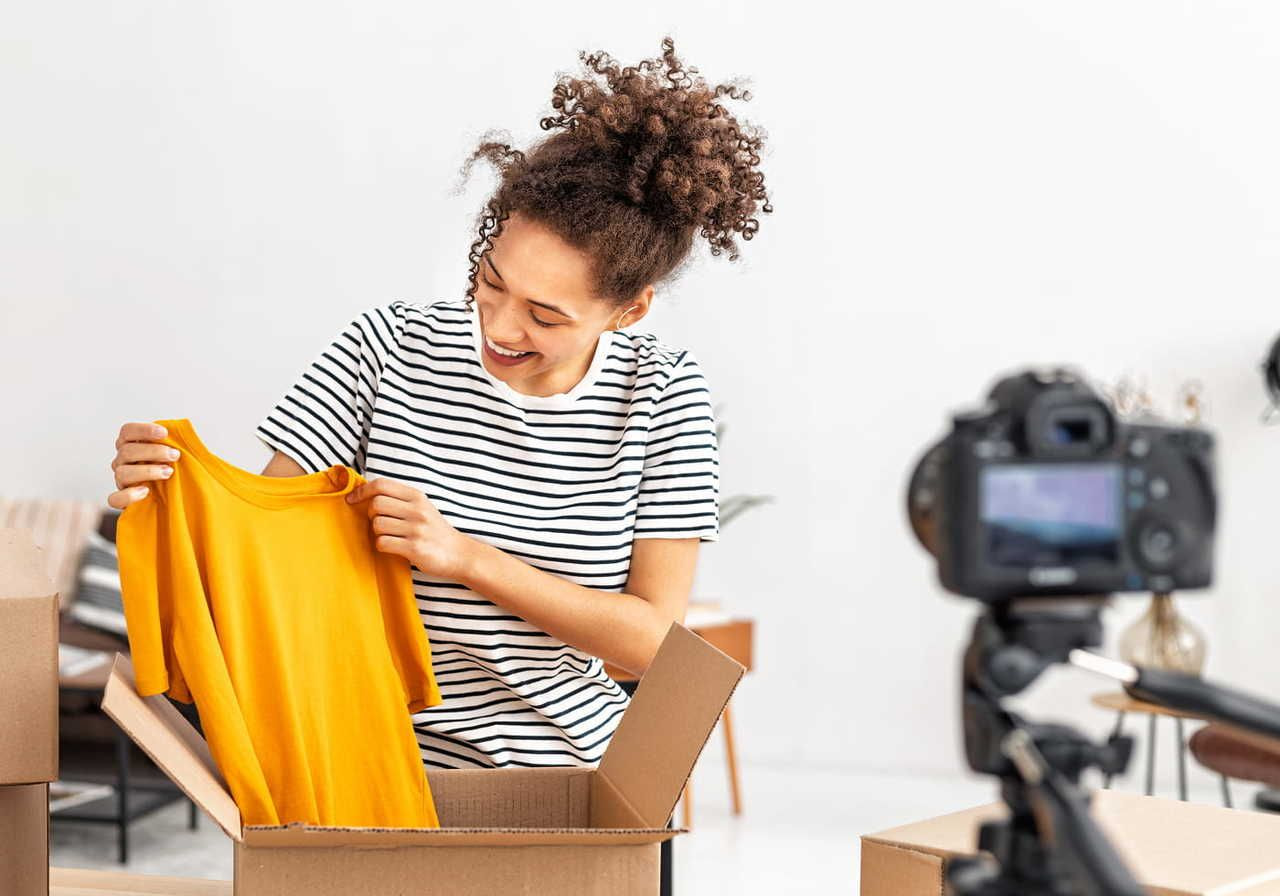 A woman with curly hair, wearing a striped shirt, holds a yellow shirt in front of a DSLR camera on a tripod.