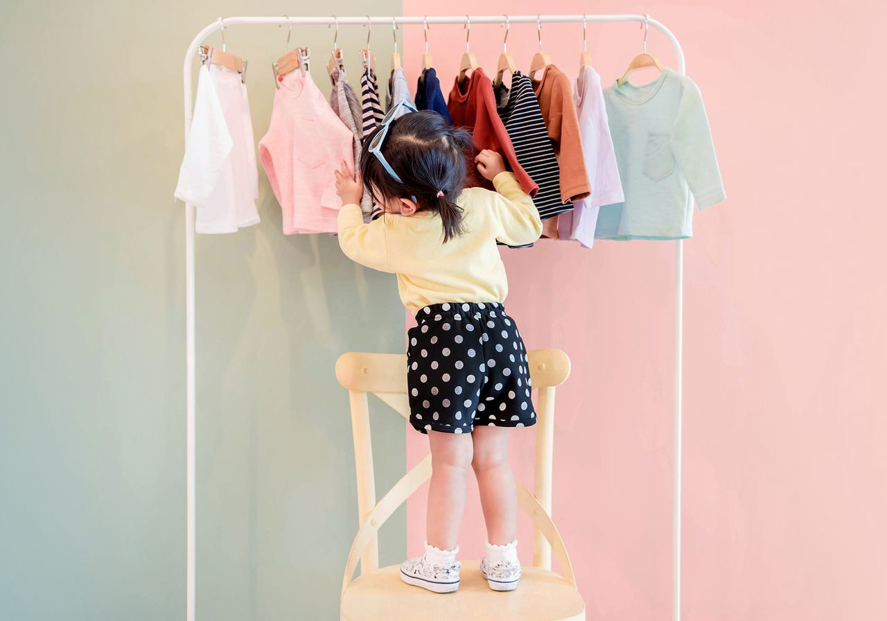 Toddler standing on a chair reaching up to browse a rack of kids' clothing.