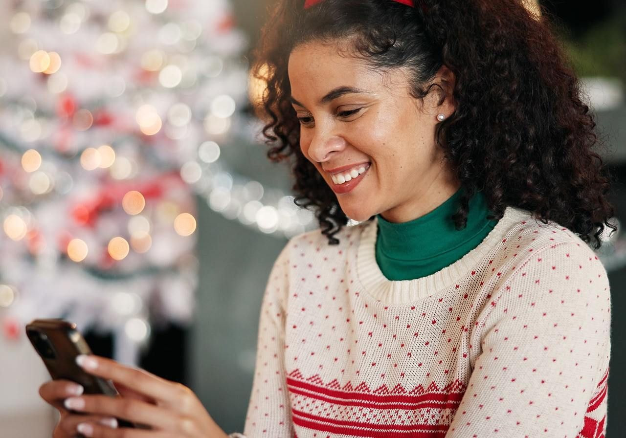 A woman in a festive Christmas sweater smiles while using her phone, radiating holiday cheer.