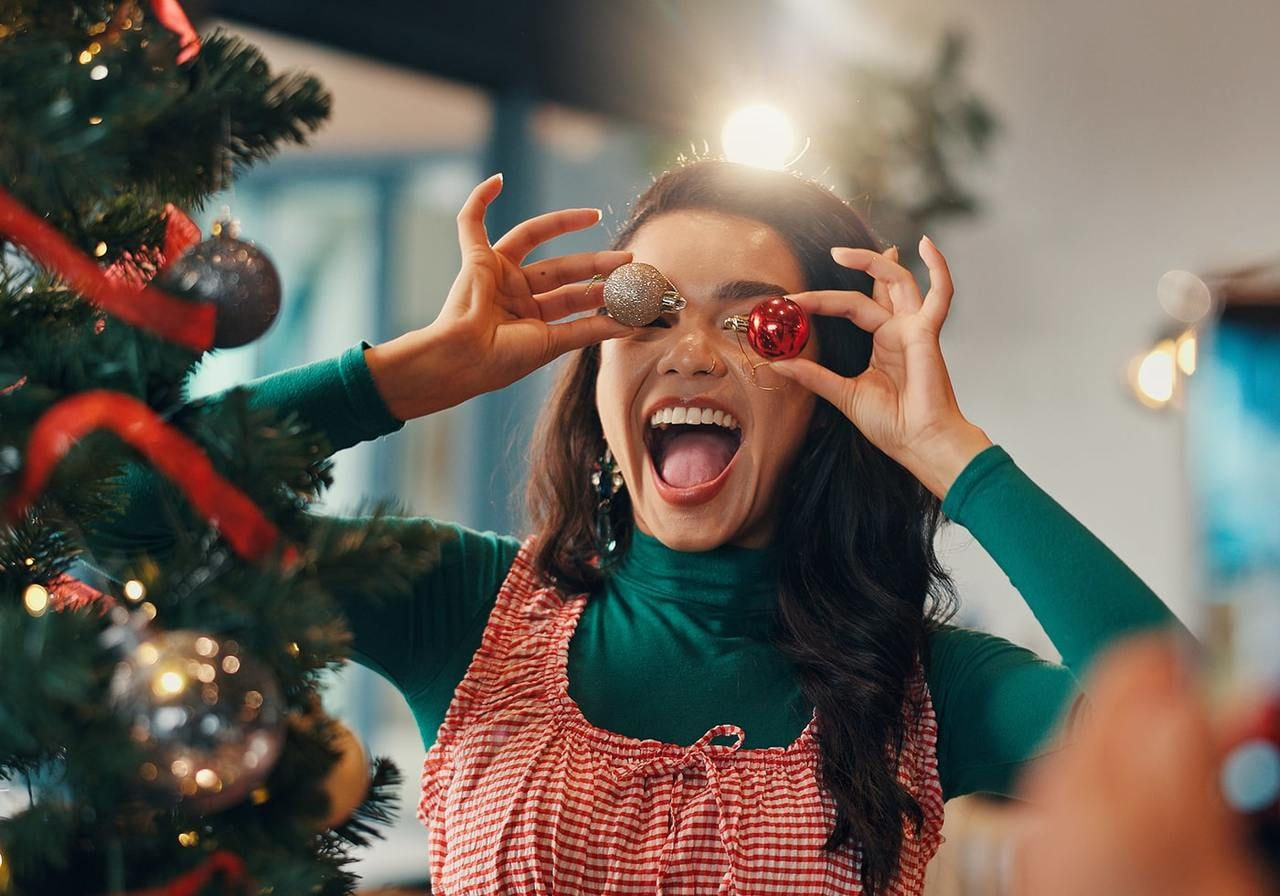 A woman holds two colorful Christmas ornaments, smiling as she prepares for the holiday season.