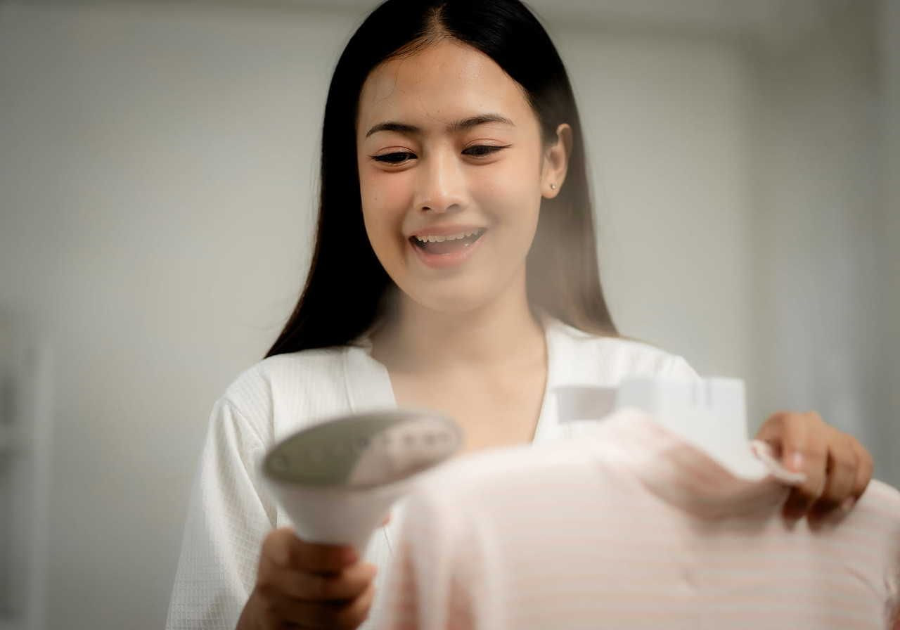 A woman is using a garment steamer to remove wrinkles from a shirt while smiling.
