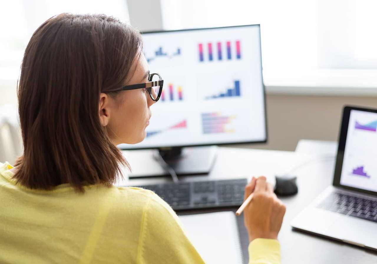 A woman with short brown hair and glasses is seated at a desk, looking at two computer screens displaying various charts and graphs.
