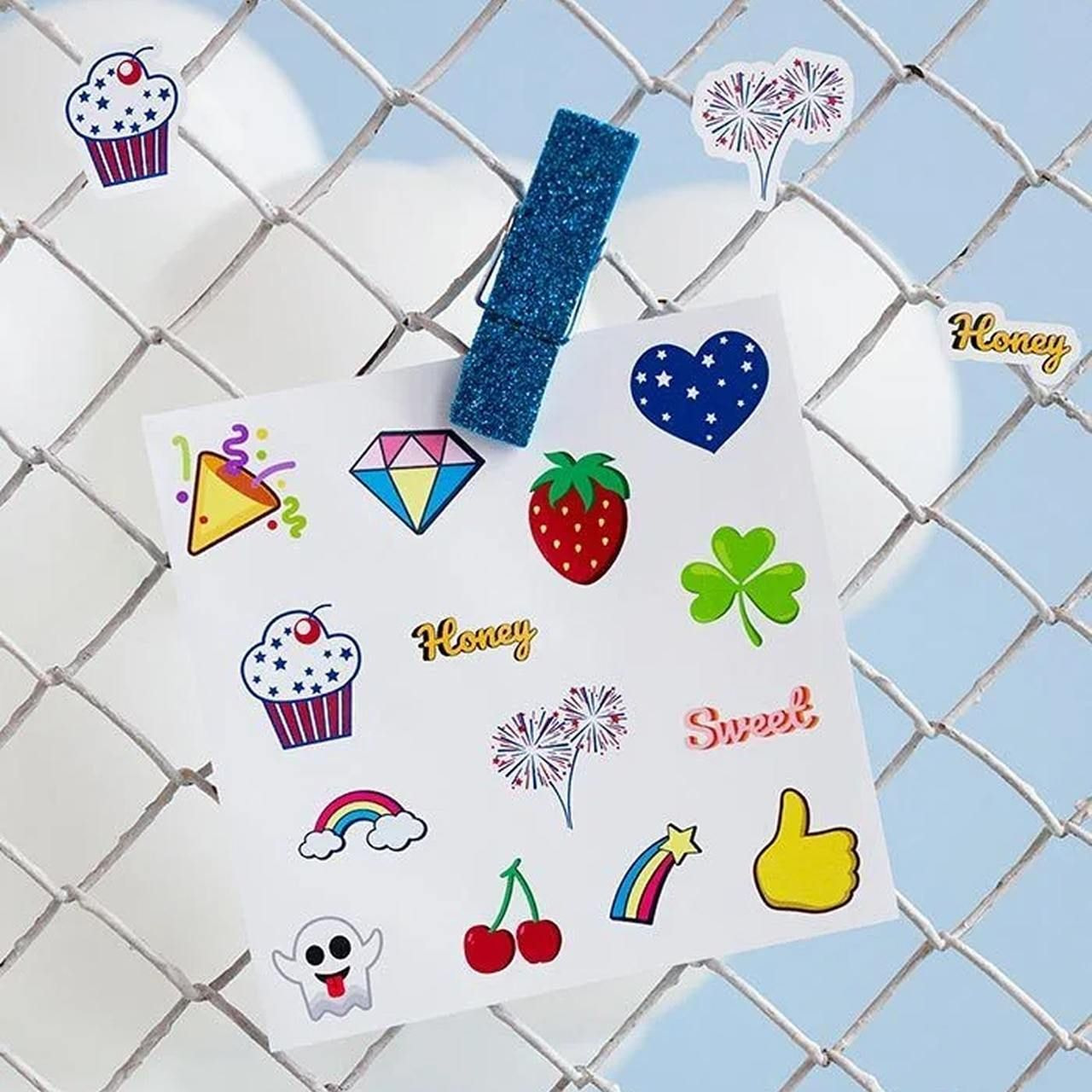 A white paper with colorful fruit-themed stickers is clipped to a wire fence using a blue glitter clip.