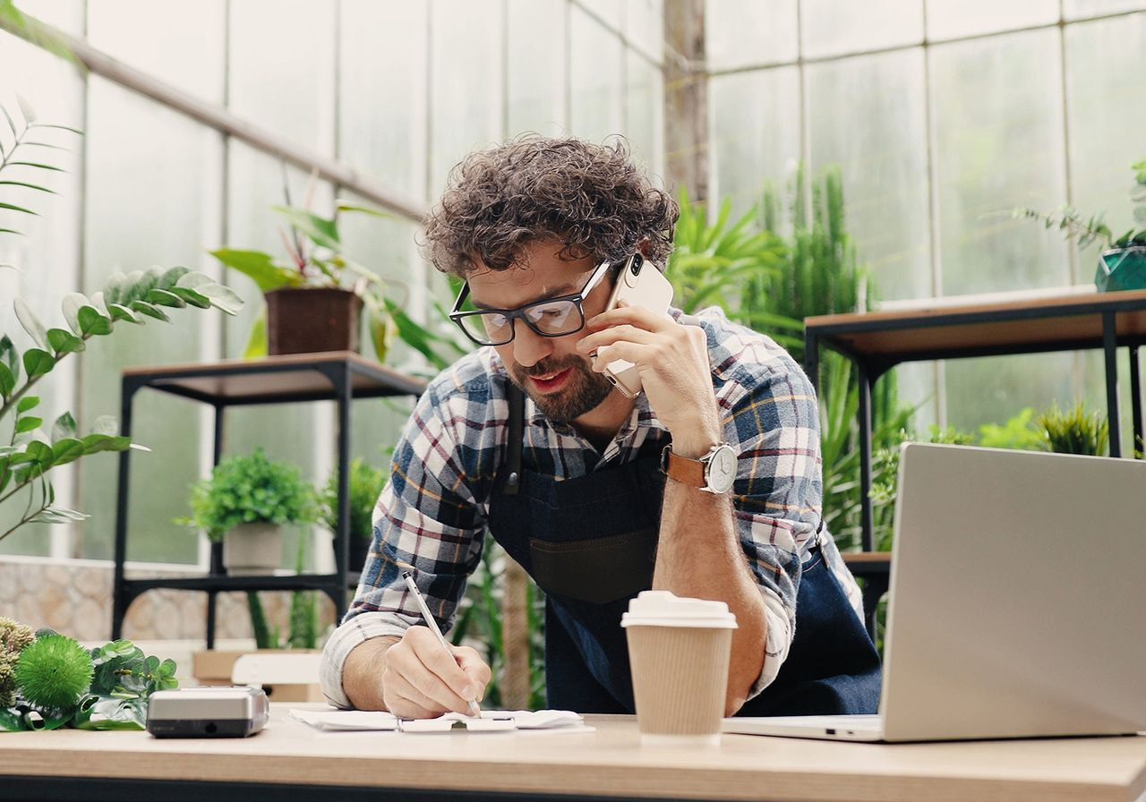 A man in an apron talks on the phone while working on a laptop at a desk, focused on his tasks.