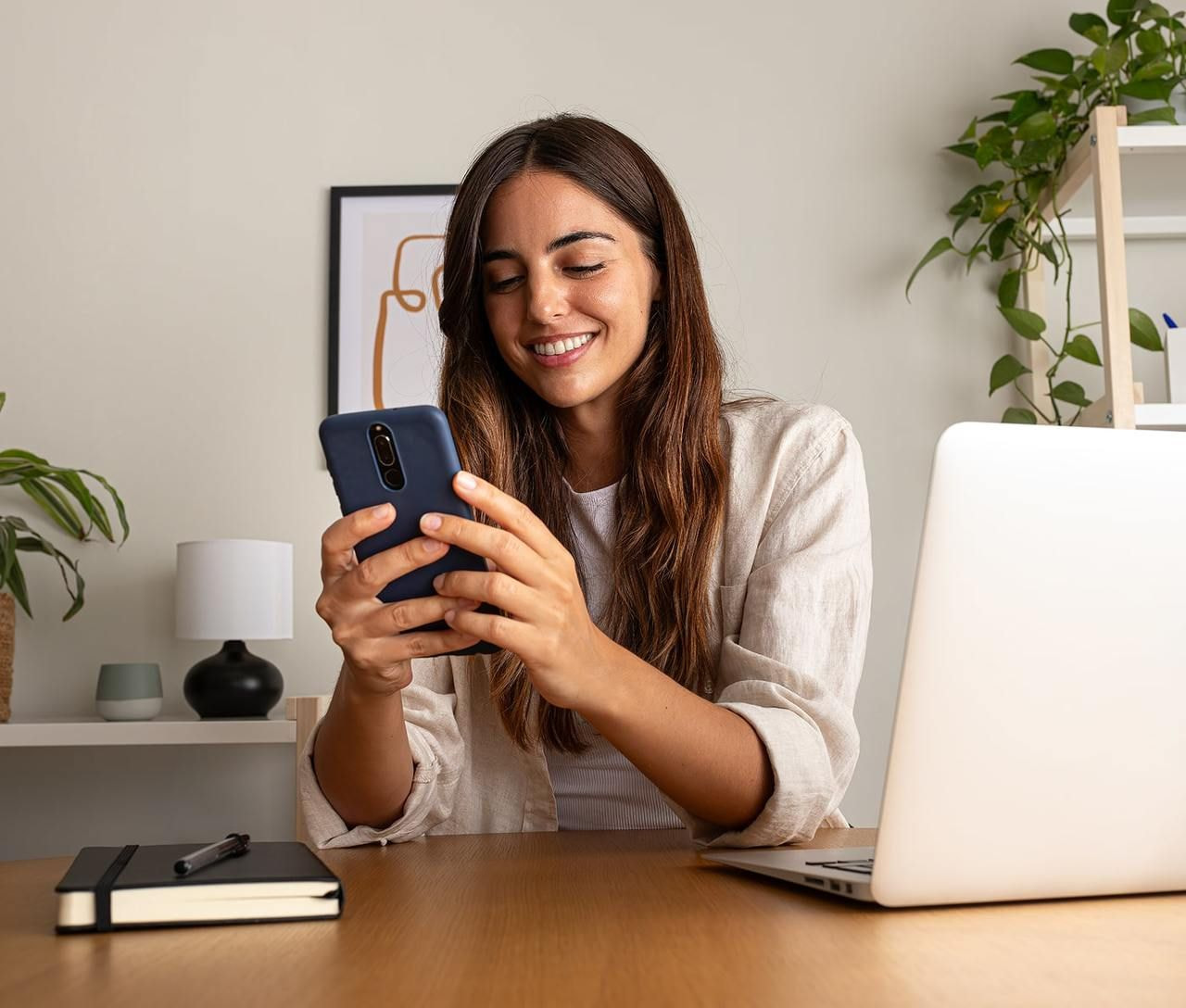 A smiling woman with long hair looks at her phone in a cozy workspace. A laptop, notebook, and potted plants are on the desk.