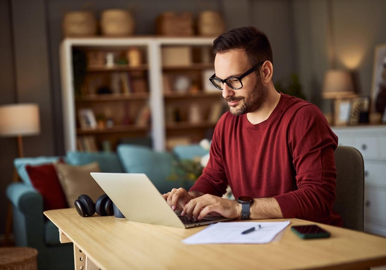 A man in a red sweater is working on a laptop at a wooden desk at home, searching for Pantone’s color of the year 2026.