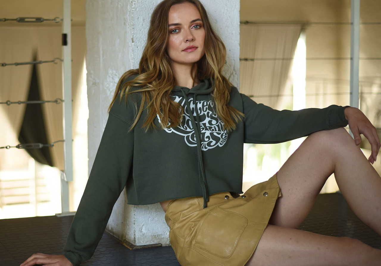 A woman sitting against a wall, wearing a cropped hoodie and a beige skirt.