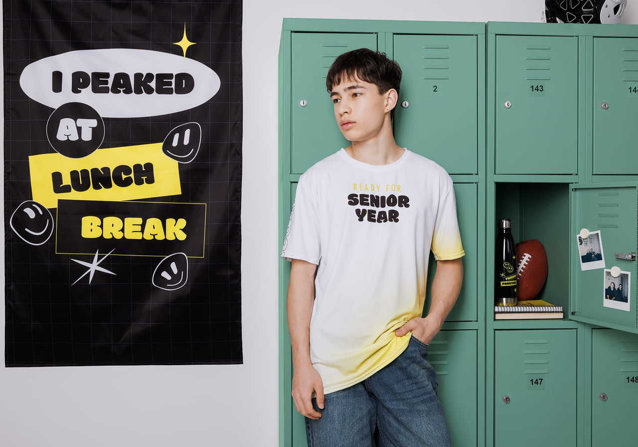 A young man in a white and yellow gradient t-shirt stands next to green lockers, with a poster that says "I peaked at lunch break" in the background.