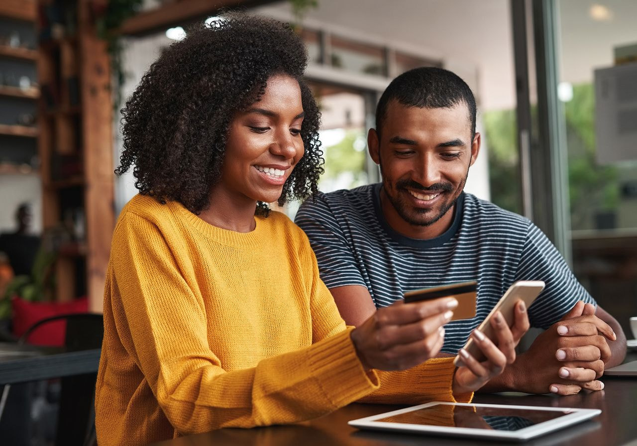 A smiling couple at a café table holds a credit card and smartphone to pay for an online order, with a tablet in front of them.