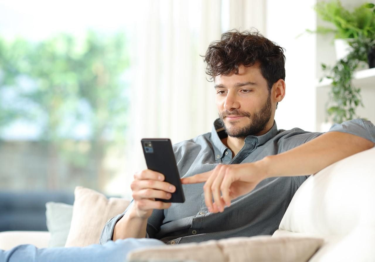 A man with curly hair relaxes on a sofa, using a smartphone. He’s focused on finding out how to start a TikTok Shop.