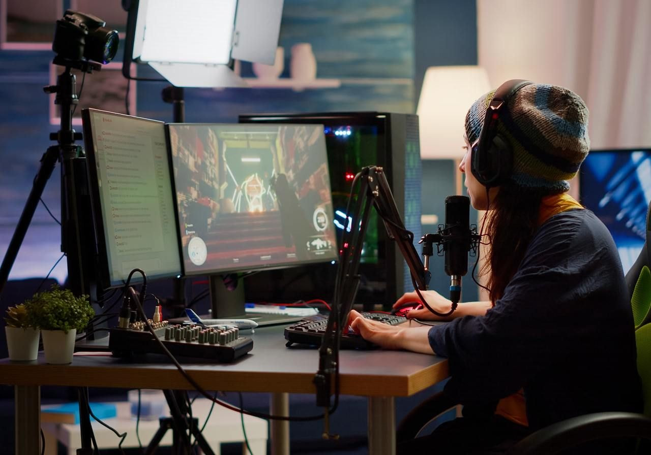 A woman wearing a beanie streams a video game at a desk with dual monitors and a microphone setup.