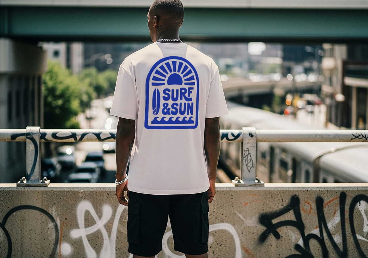 A man in a "Surf & Sun" t-shirt faces away on an urban bridge. The shirt has a blue sun and waves graphic.