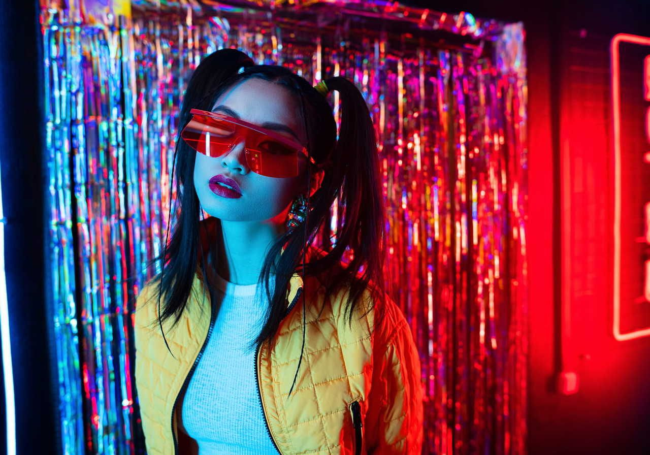 A young woman with pigtails wears red sunglasses and a yellow jacket, posing against a colorful, metallic backdrop with neon lights.