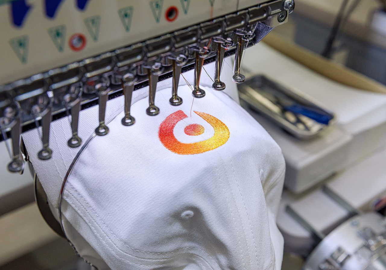 A white hat with an embroidered logo featuring a circular design in orange and yellow, placed on an embroidery machine.