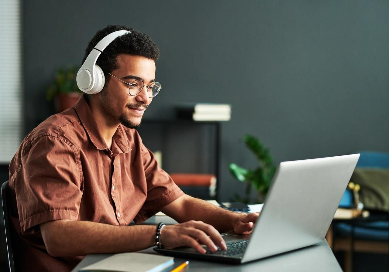 A young man in glasses and wearing headphones is using a laptop.