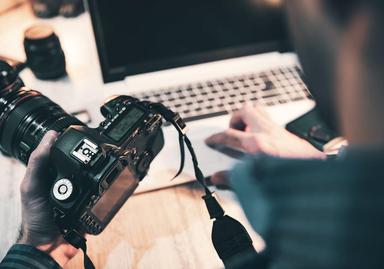 A person holding a camera in one hand and a laptop in the other, standing against a neutral background.