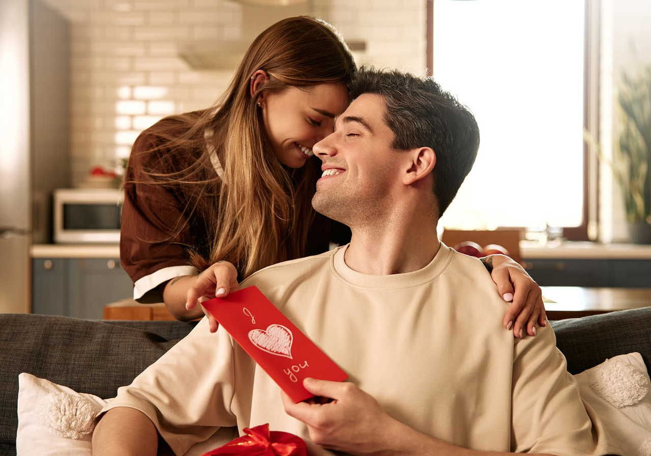 A woman leans affectionately over a smiling man holding a red card with "I love you" written on it.