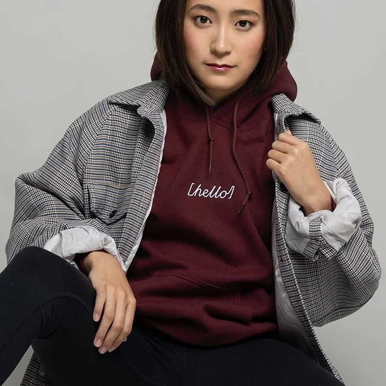 A woman wearing a burgundy hoodie with the word "hello" and a checkered jacket. She has a calm expression, sitting against a plain background.