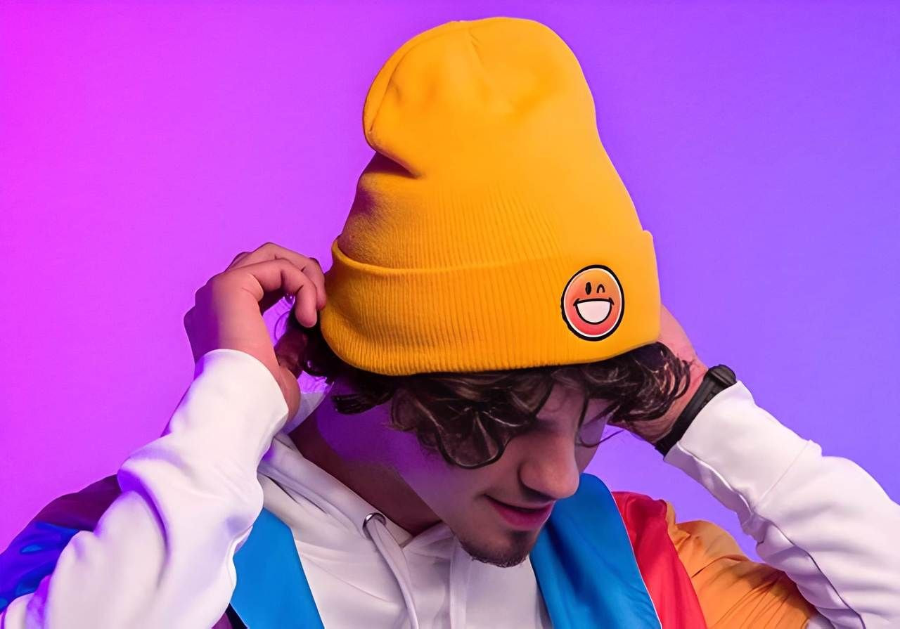 A man wearing a bright yellow beanie with a smiley face adjusts it. He’s dressed in a colorful hoodie. The background is a vibrant purple.