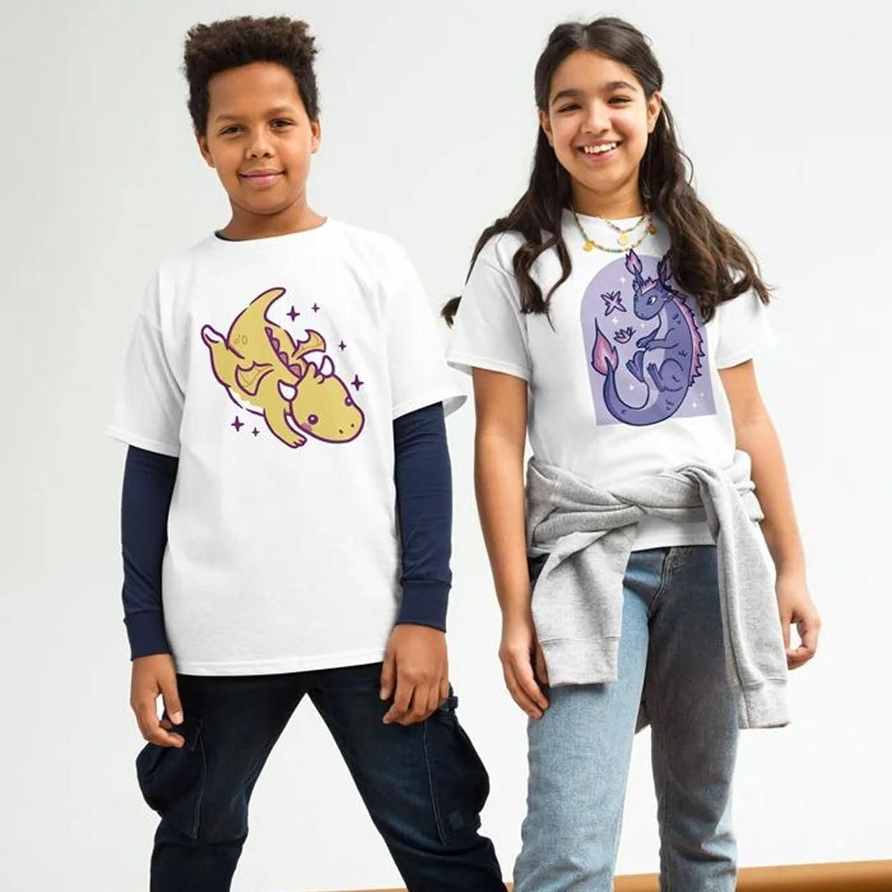 Two children smile, wearing white shirts with cartoon creatures. The boy's shirt has a yellow dinosaur, the girl's a purple dragon.