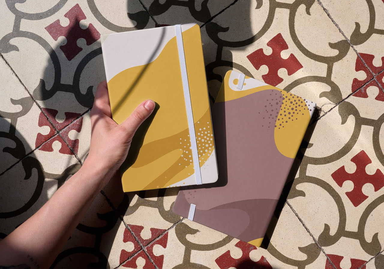 A person holding two reading journals with abstract designs, as an example of a gift idea for a book lover, on a patterned floor.