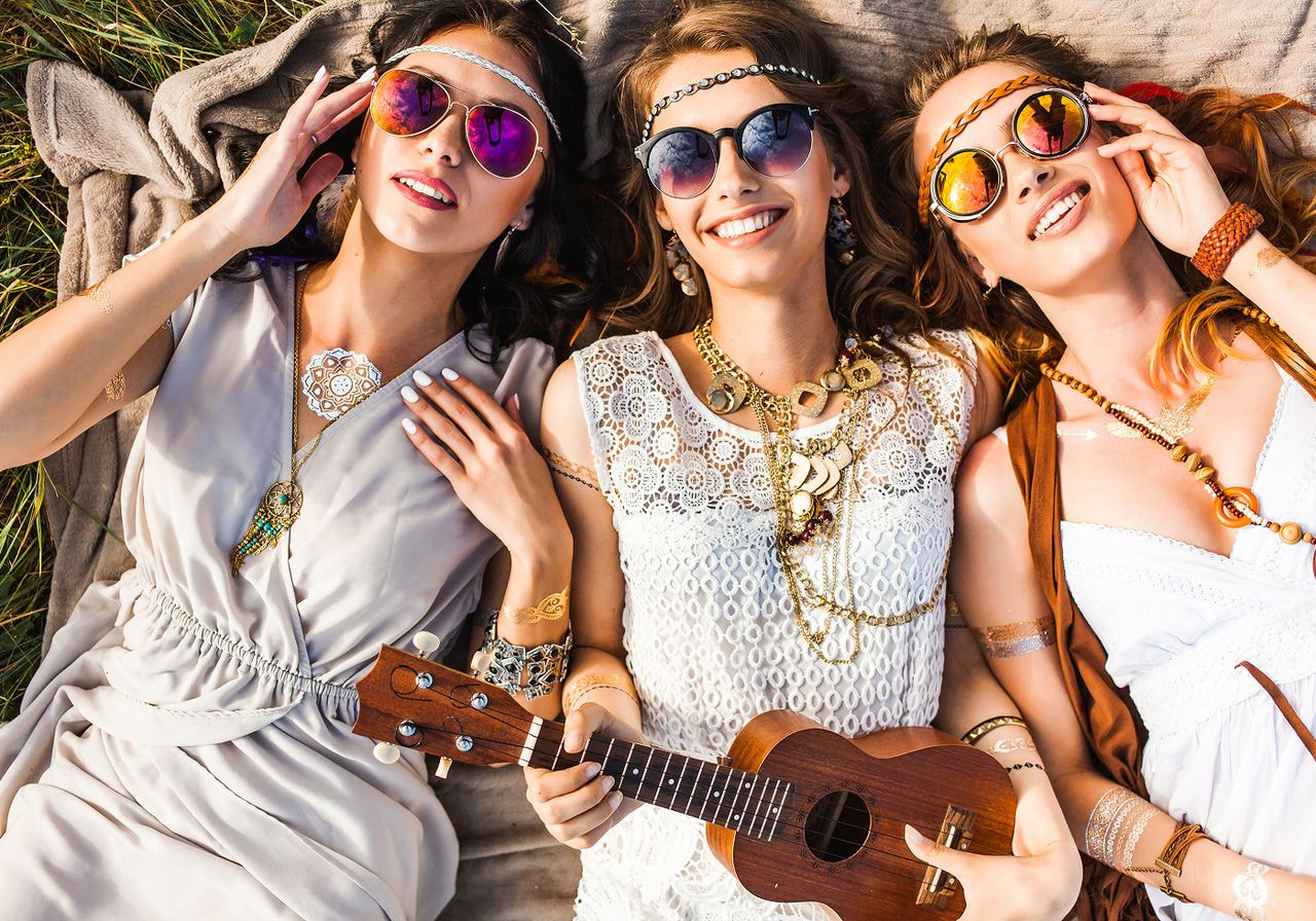 Three women lie on a blanket in a field, smiling and wearing bohemian outfits, jewelry, and sunglasses.