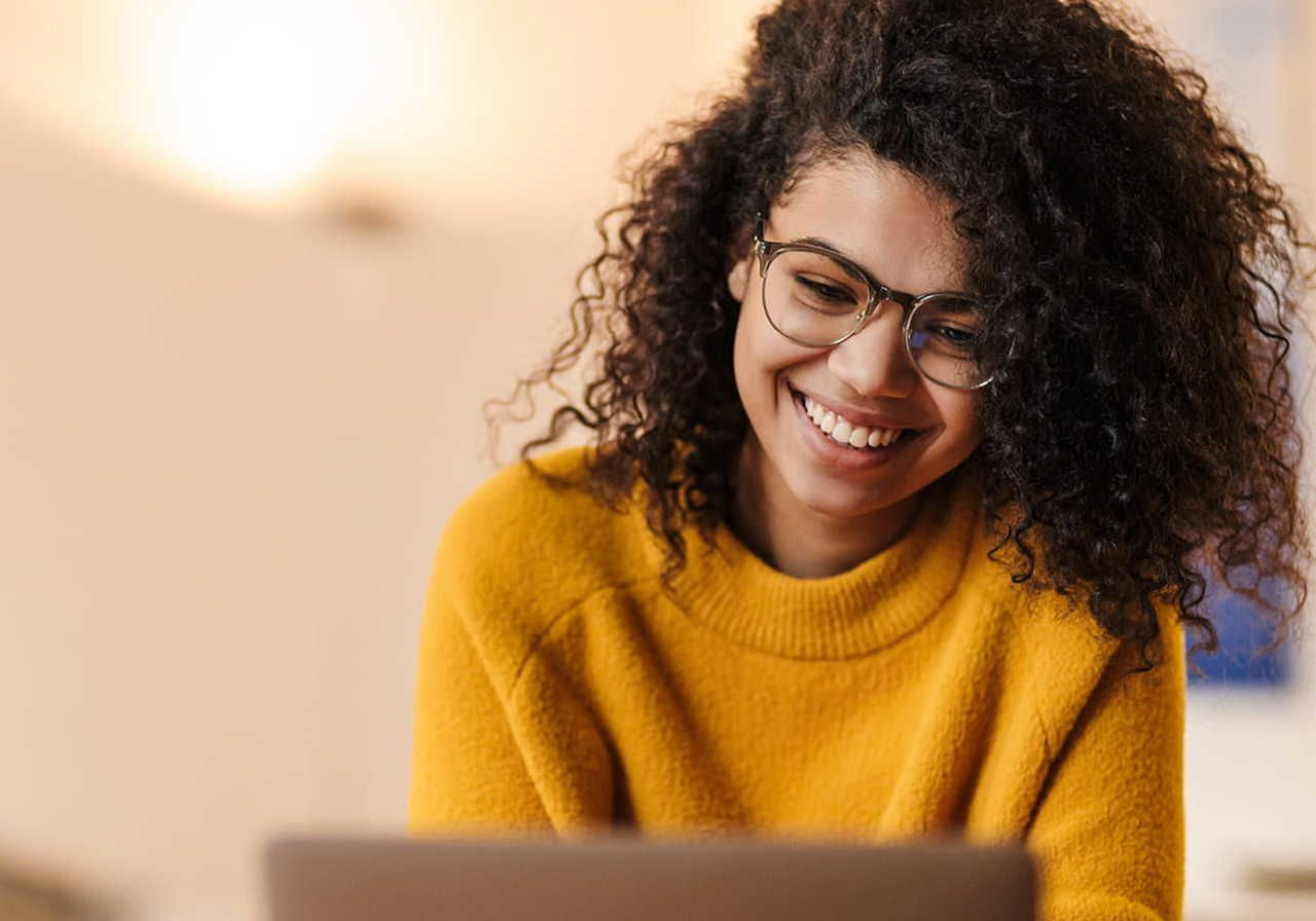 A smiling woman with curly hair and glasses in a yellow sweater sits at a desk, using a laptop to learn how to start a sustainable clothing brand.