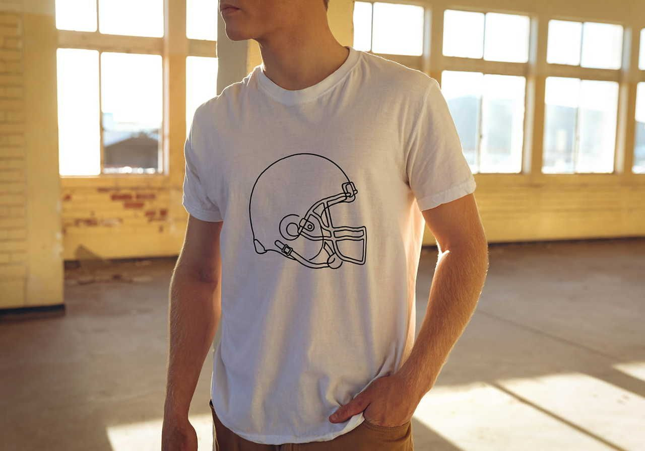 Man in a white t-shirt featuring a football helmet outline, standing in a sunlit room with large windows.