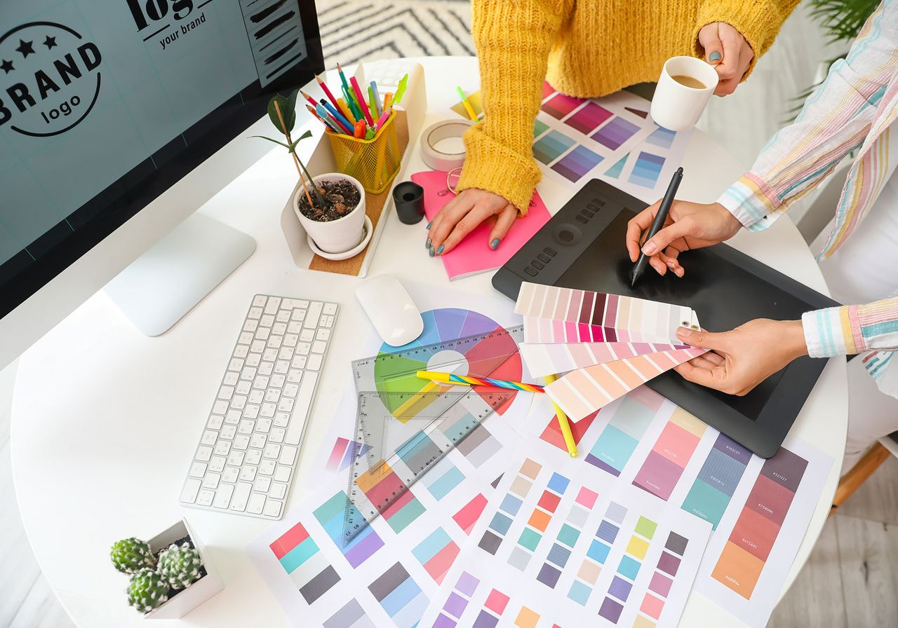 Two designers reviewing color swatches and a logo mockup on a computer screen at a desk.