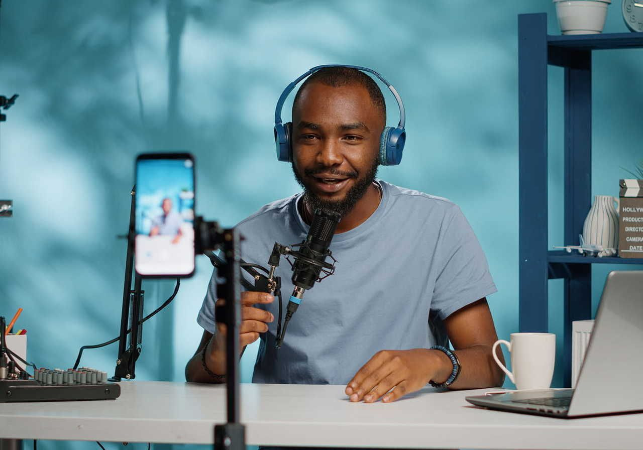 A person with headphones and a microphone records a podcast in a studio, smiling at a smartphone camera.