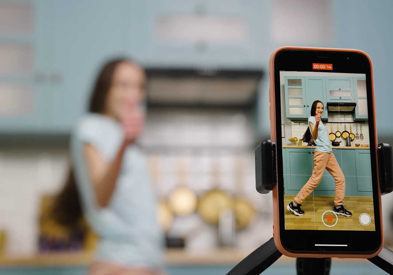 A smartphone on a tripod records a person dancing in a kitchen. The phone screen shows the dancer clearly.