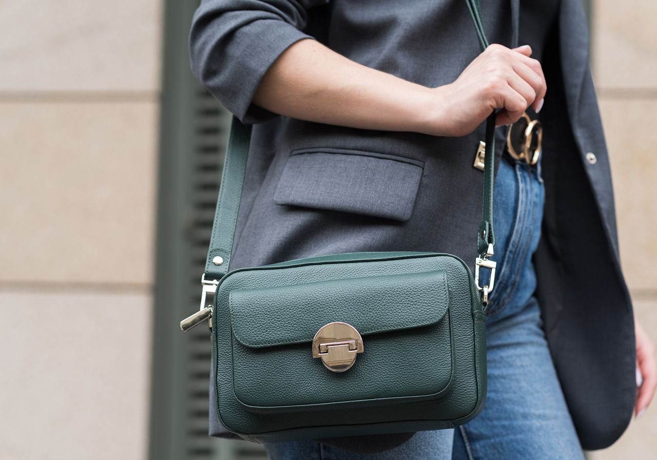 A woman wearing jeans and a blazer holds a stylish green leather bag, standing confidently.