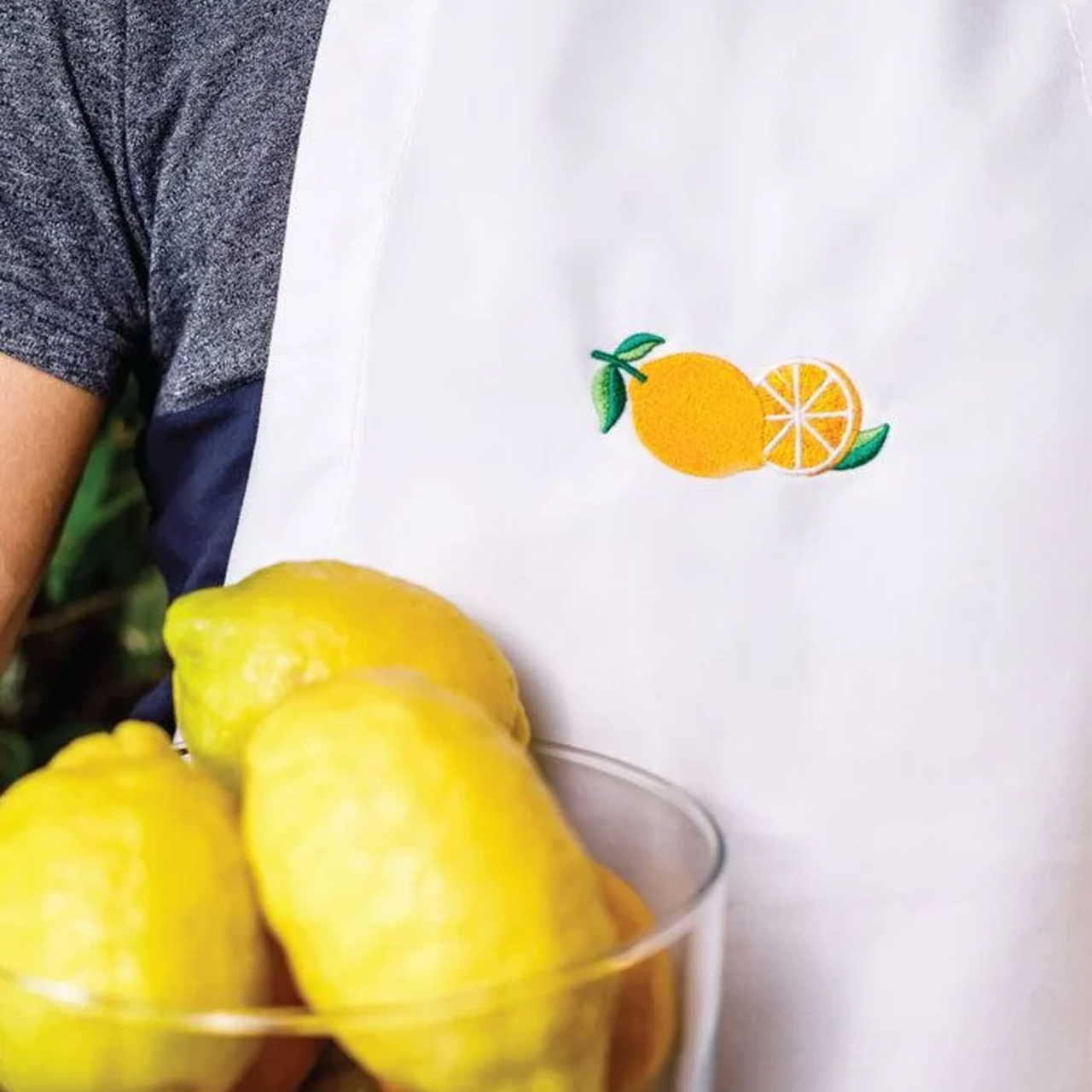 A person in a blue shirt wears a white apron with lemon embroidery, holding a glass bowl of bright yellow lemons.