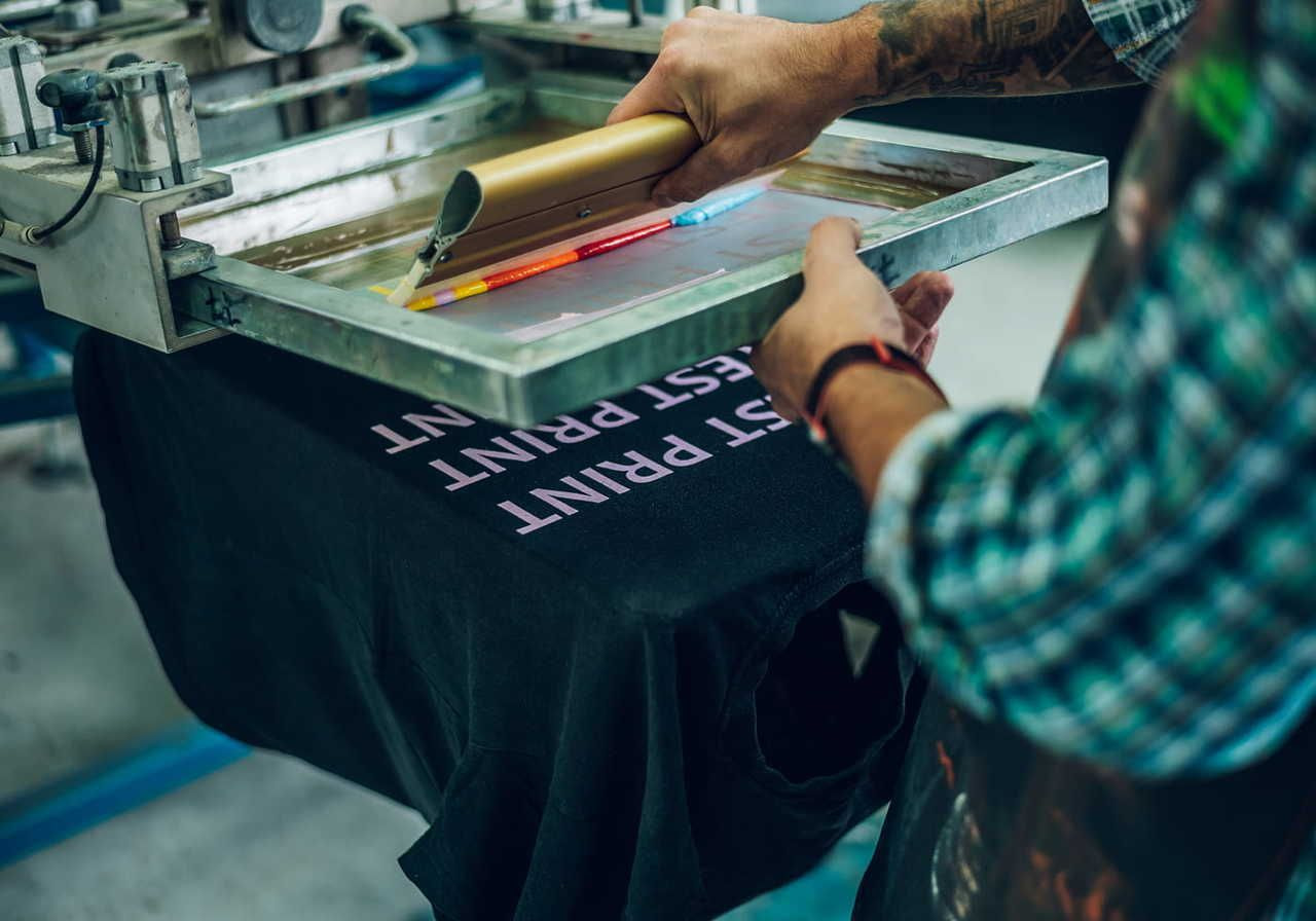 A person in a plaid shirt uses a squeegee on a screen printing frame to print white text on a black t-shirt.