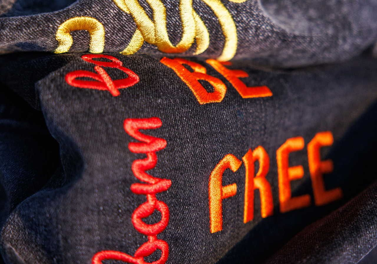 Close-up of a dark fabric with embroidered text that reads "Born to be FREE" in orange and yellow thread.