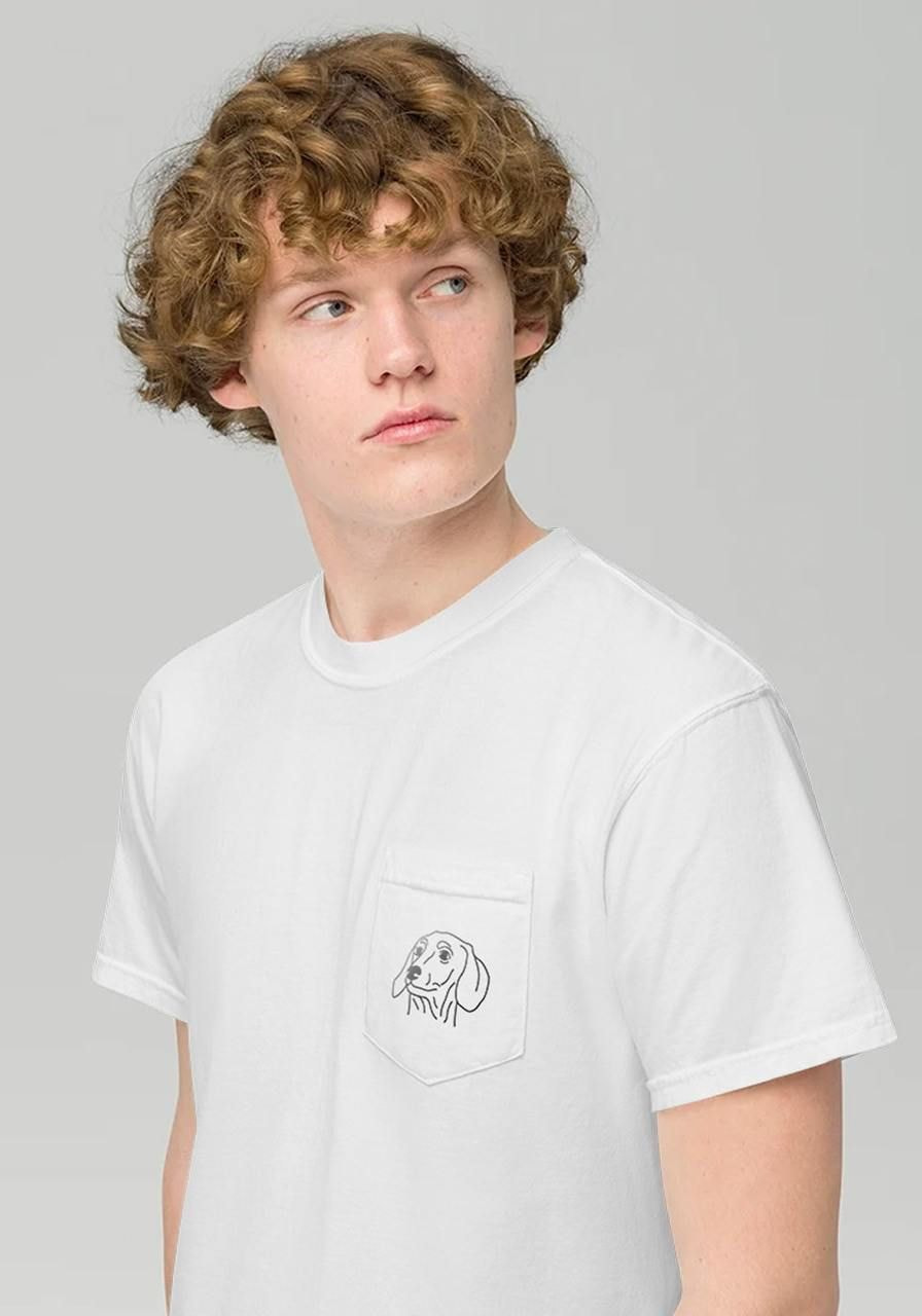 A young man with curly hair in a white t-shirt looks to the side, with a t-shirt that has a dachshund print on the pocket.