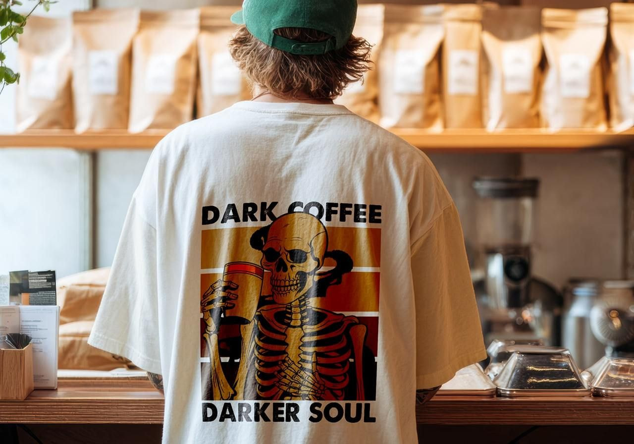 A person in a café wearing a t-shirt featuring a skeleton holding coffee with the text "Dark Coffee, Darker Soul."