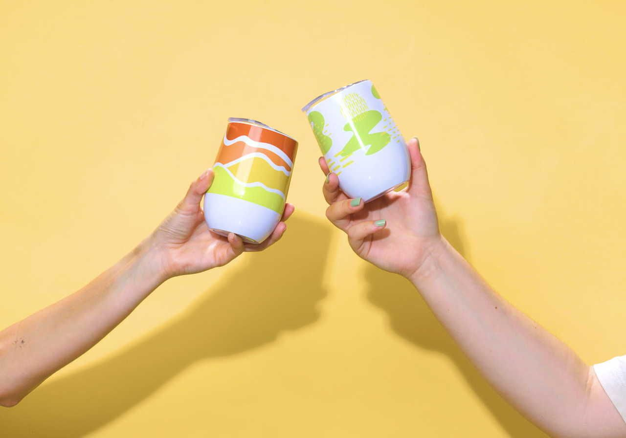 Two hands hold colorful, patterned custom wine tumblers, clinking them against a bright yellow background.