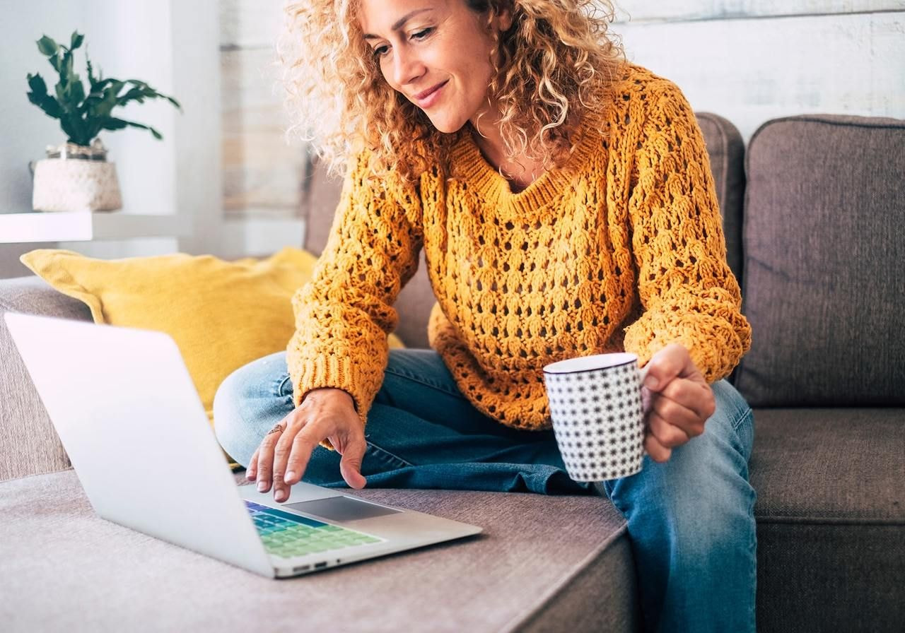 A woman in a cozy yellow sweater sits on a gray sofa, smiling at a laptop, holding a coffee mug.