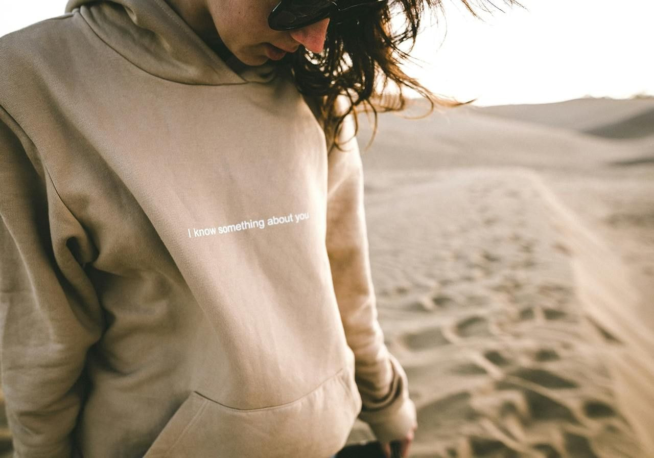 A woman in a cream “I know something about you” hoodie in a sand dune location. 