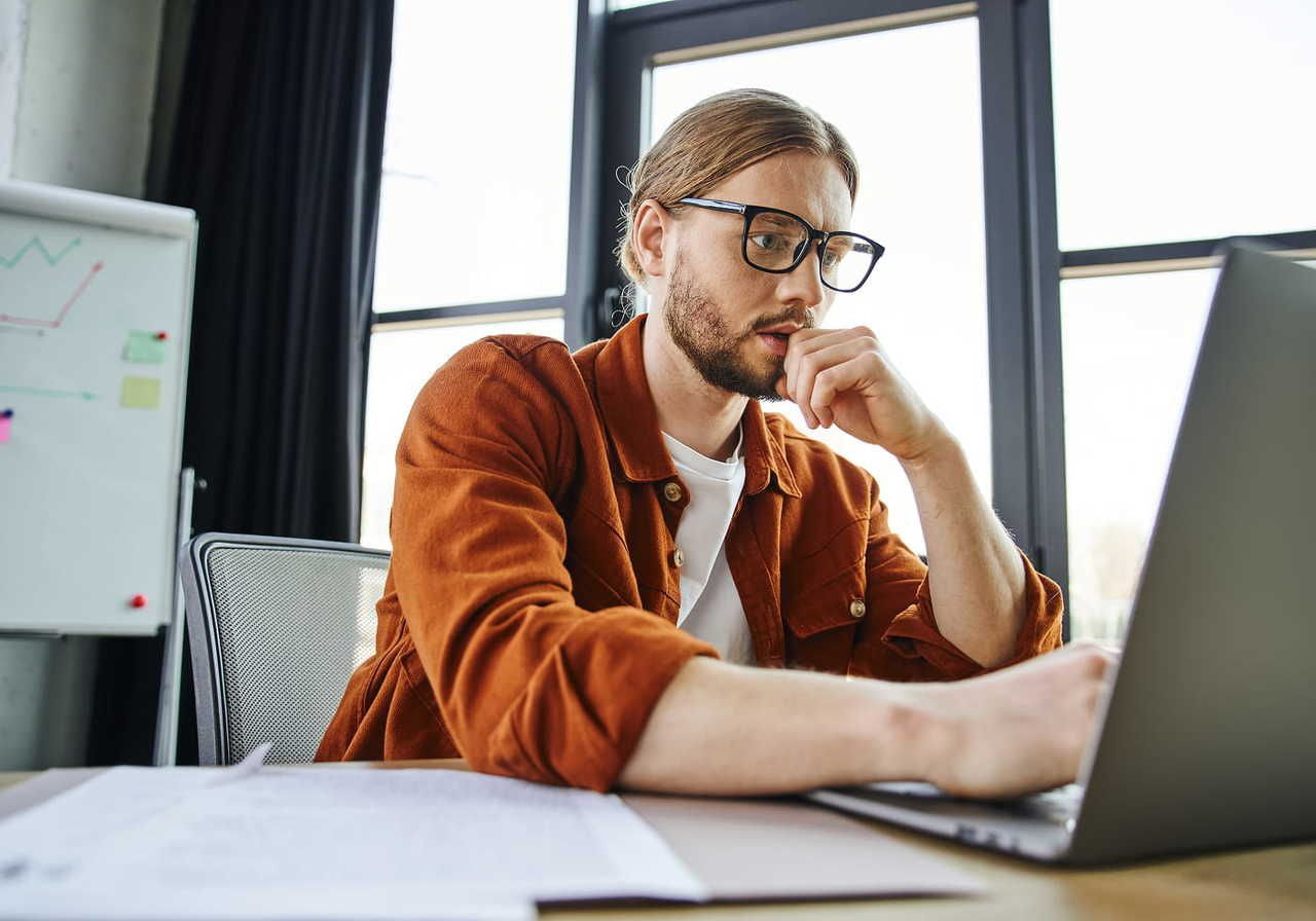 A man with glasses and a beard, wearing a brown jacket, intensely focuses on a laptop. Behind him is a whiteboard with graphs.
