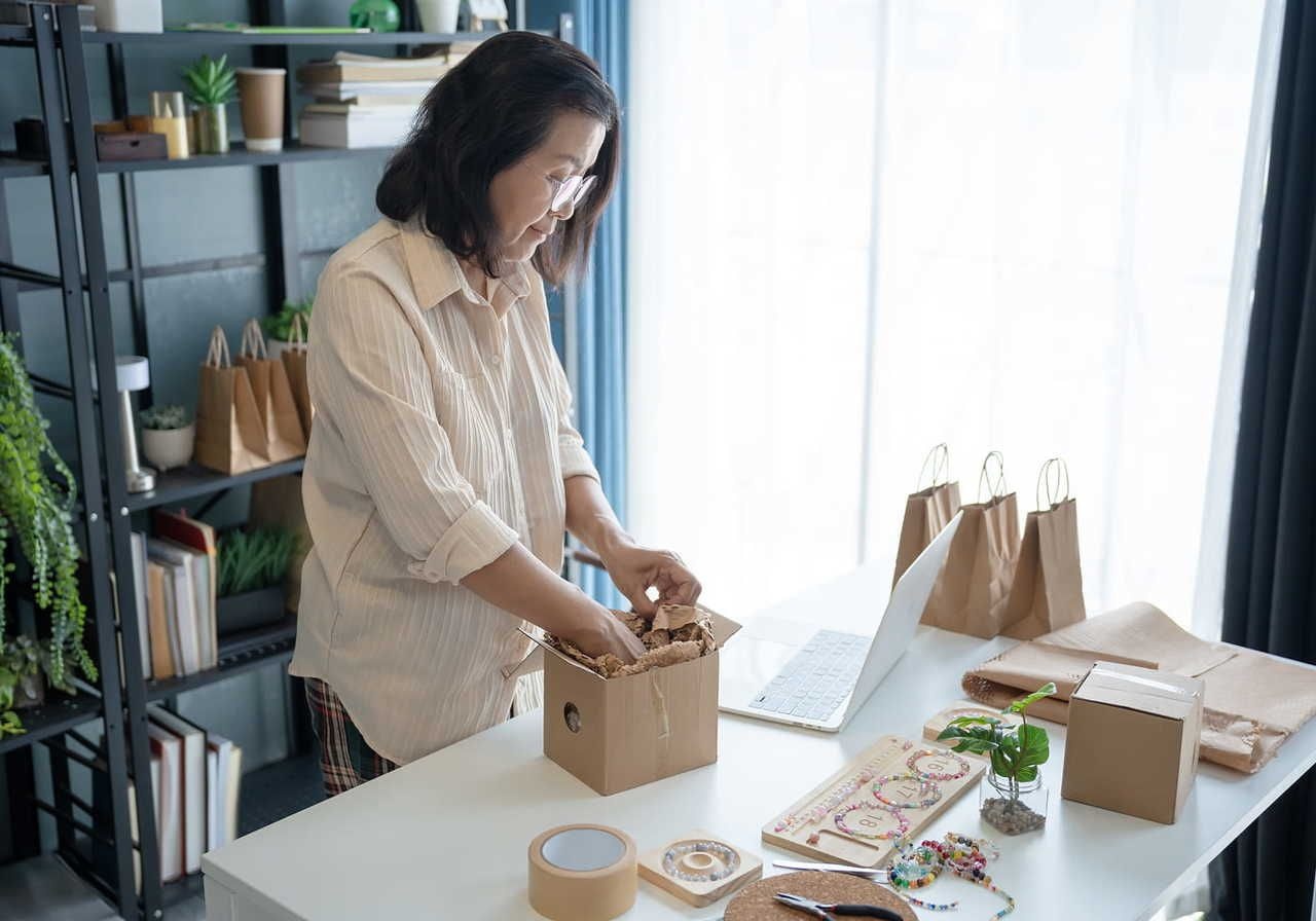 A woman in a light blouse is packing a product into a cardboard box, in what seems to be a home office.
