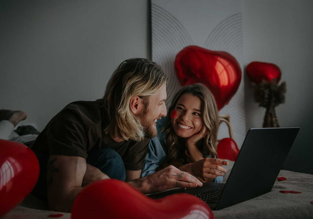 A couple lies on a bed, smiling at each other, with a laptop in front. Red heart-shaped balloons surround them.