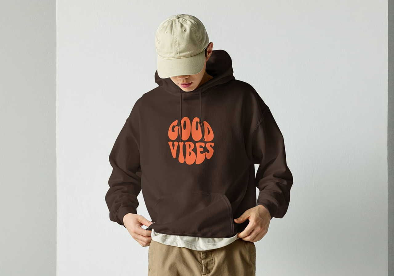 A man wearing a beige cap and a brown hoodie with "Good Vibes" in bold orange text. He’s gazing downward, adjusting the hem.