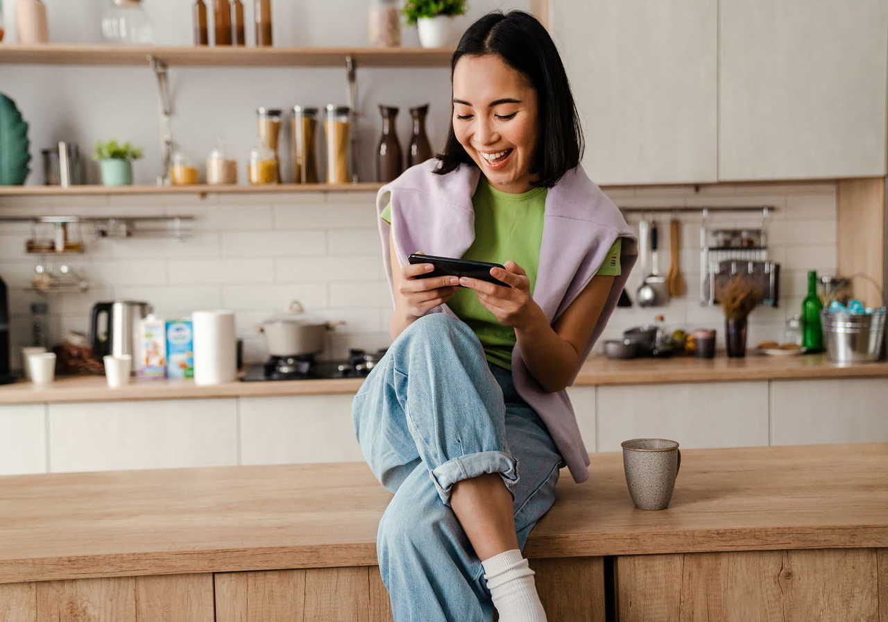 A young woman sits on a kitchen counter, smiling at her phone. She's wearing casual clothing.