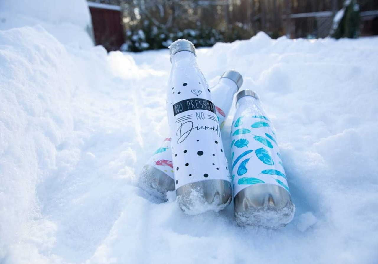 Three water bottles in the snow, one white with black dots and text, another white with blue designs, and the third white with black text.