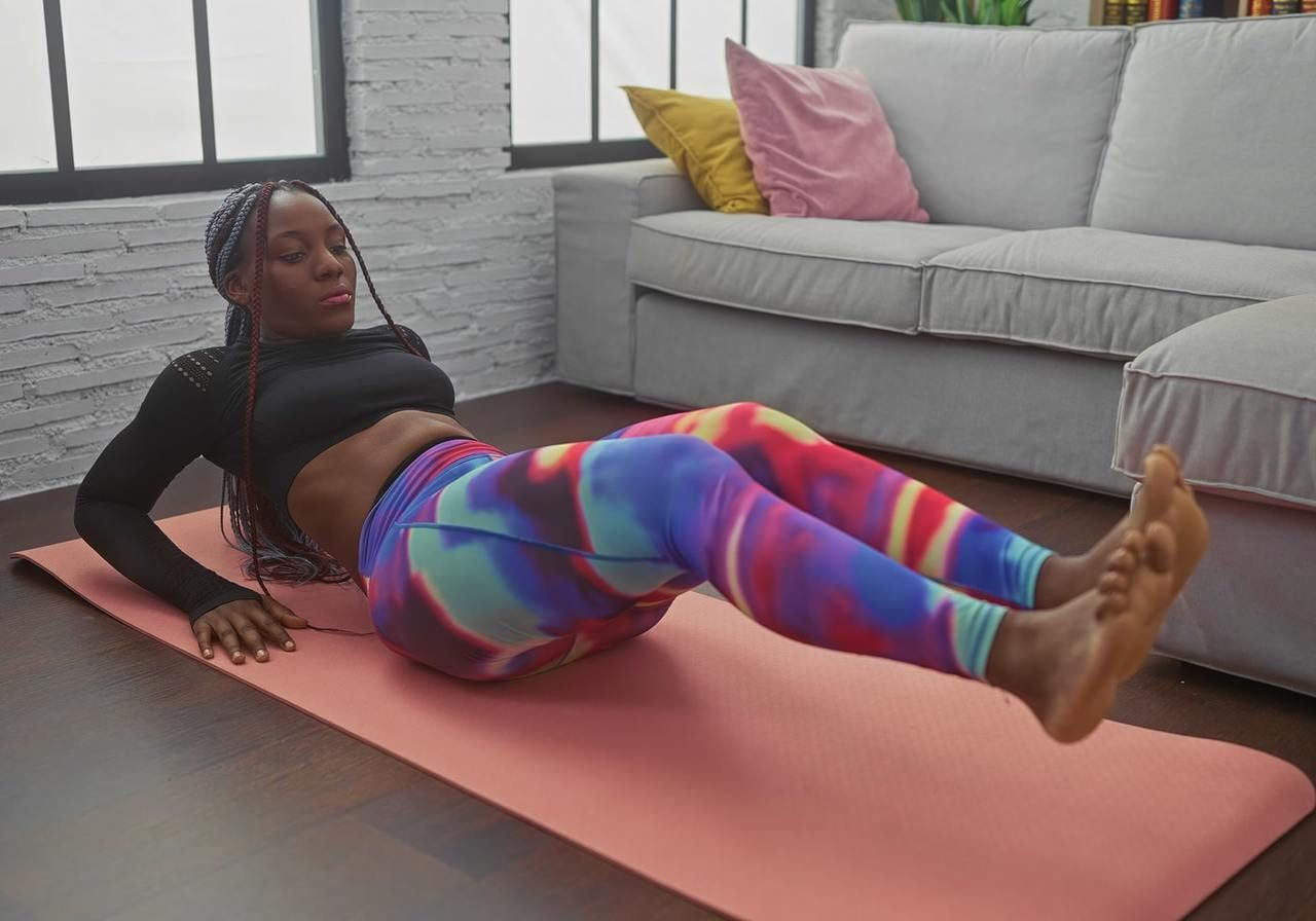 A woman exercising on a pink yoga mat indoors, performing a leg raise. She wears a black top and vibrant multi-colored leggings.