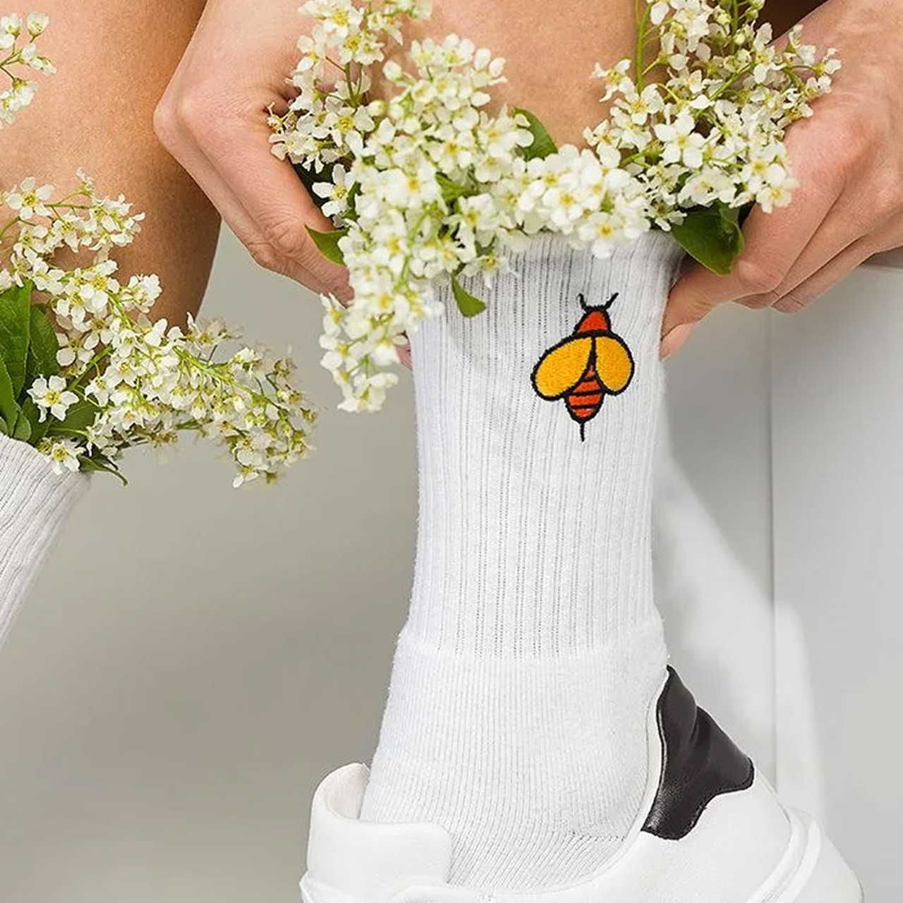 White sock with bee embroidery and flowers tucked inside, worn with white sneakers. Hands gently holding the flowers.