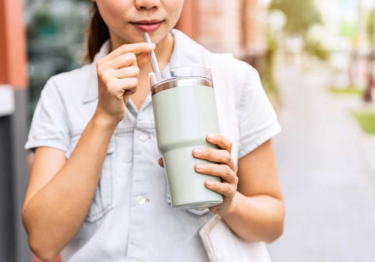 A woman in a sleeveless shirt drinks from a large, light green tumbler with a straw. She stands on a sunlit street.